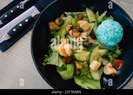 Blauer Reis und Salat mit Gemüse und Meeresfrüchten. Grüner Salat in dunkler Schüssel mit Besteck. Thailändische Küche. Gesunde Ernährung. Asiatische Mahlzeit. Gesunder Lebensstil. Stockfoto