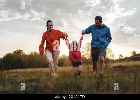 Die Familie genießt einen spielerischen Lauf auf dem Feld bei Sonnenuntergang, mit einem Kind, das zwischen den Eltern schwingt und die Freude am aktiven Familienleben und die Schönheit von n verkörpert Stockfoto