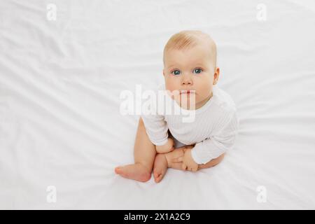 Das charmante kleine Mädchen in kuscheliger weißer Kleidung sitzt bequem auf dem weichen Bett und bietet einen neugierigen Blick direkt in die Kamera. Stockfoto