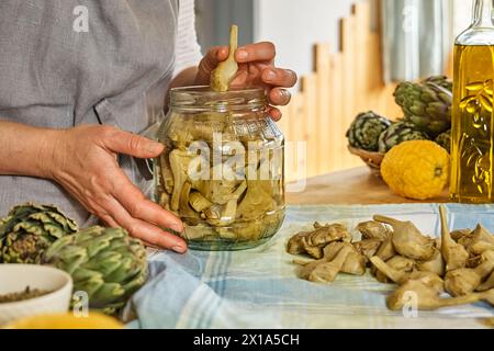 Frau, die italienische Artischocken in Olivenöl zubereitet. Artischockenherzen eingelegt mit Olivenöl und Kräutern. Hausgemachte gesunde Ernährung. Stockfoto