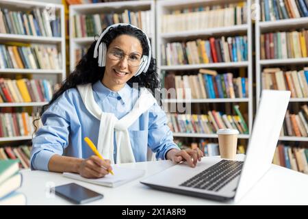 Fröhliche Frau mit Kopfhörern, die am Büchertisch studiert, umgeben von Büchern. Sie schreibt Notizen und benutzt einen Laptop, der eine konzentrierte und entspannte Atmosphäre vermittelt. Stockfoto