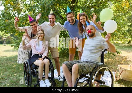 Eine Gruppe von Freunden, einschließlich Rollstuhlfahrer, genießen eine lebhafte Outdoor-Party mit Ballons, bei der Inklusion und Freude in einem üppigen Park gefeiert werden Stockfoto