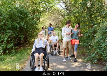 Eine vielfältige Gruppe von Freunden, darunter Rollstuhlfahrer, teilen Lachen und Gespräche während eines gemütlichen Spaziergangs in einem üppigen Park. Stockfoto