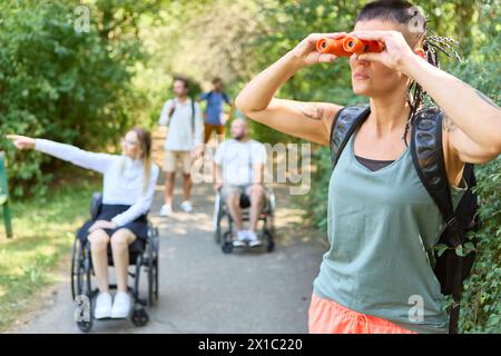 Outdoor-Szene mit verschiedenen Freunden, mit Fokus auf Einbeziehung, da Frau im Rollstuhl interagiert und ein anderer mit Fernglas. Stockfoto