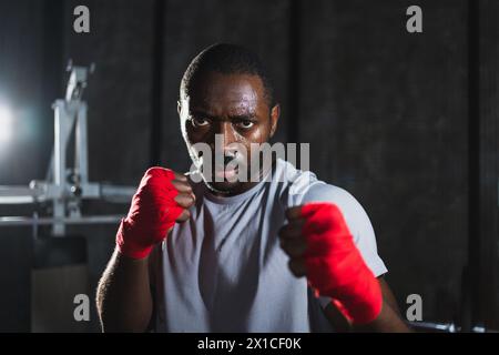 Boxer im Fitnessstudio. Aggressiver Afrikanerkämpfer, der sich nach dem Training im Boxen ausruht, bereit für den Kampf, mit Blick auf die Kamera. Stark geschwitzter Mann, der Stanzungen ausübt, sieht konzentriert aus und bereitet sich auf den Sparring vor Stockfoto