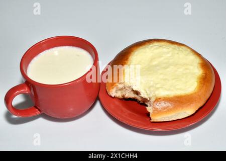 Auf dem Tisch steht eine braune Tasse Milch und ein Brötchen, ein Käsekuchen mit abgebissenem Hüttenkäse auf einer Seite, auf einem Teller. Stockfoto