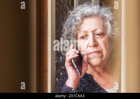 Nahaufnahme einer 70-jährigen Frau, die von einem Fenster seines Hauses aus auf dem Handy spricht, während sie nach draußen blickt. Stockfoto
