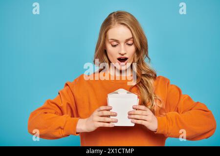 Eine atemberaubende blonde Frau in einem orangefarbenen Pullover hält eine weiße Lebensmittelbox Stockfoto