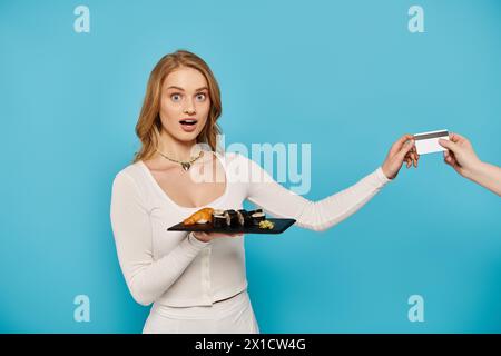 Eine schöne blonde Frau in einem weißen Kleid hält elegant ein Tablett mit köstlicher asiatischer Küche, Hand mit Kreditkarte. Stockfoto