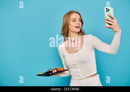 Blonde Frau, die ein Selfie mit ihrem Handy macht, während sie köstliches asiatisches Essen hält und eine Pose schlägt. Stockfoto