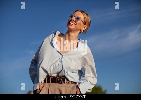 Moderne Frau in übergroßem Hemd und Sonnenbrille, fröhlich lächelnd am blauen Himmel. Trendige, fröhliche und stilvolle Sommeratmosphäre. Stockfoto