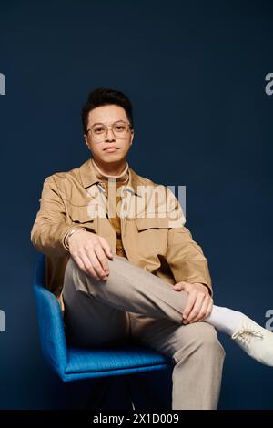 Ein modischer junger Mann in eleganter Kleidung sitzt auf einem blauen Stuhl und kreuzt seine Beine in selbstbewusster Pose. Stockfoto