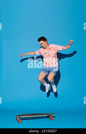 Ein stilvoller junger Mann in trendiger Kleidung springt mit einem Skateboard vor einem leuchtend blauen Hintergrund in die Luft. Stockfoto