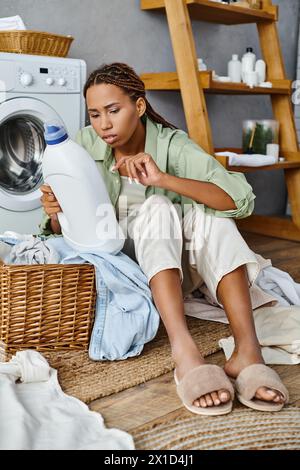 Afroamerikanerin mit Afro-Zöpfen, die Wäsche waschen, sitzt auf dem Boden neben einer Waschmaschine im Badezimmer. Stockfoto