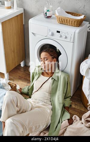 Afroamerikanerin mit Afro-Zöpfen, die neben einer Waschmaschine sitzt und im Badezimmer Haushaltswäsche macht. Stockfoto