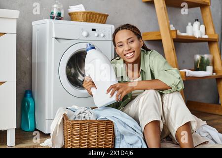 Eine Afroamerikanerin mit Afro-Flechten sitzt neben einer Waschmaschine und wäscht im Badezimmer. Stockfoto