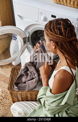 Afroamerikanerin mit Afro-Zöpfen sitzt neben einer Waschmaschine und wäscht im Badezimmer. Stockfoto