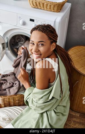 Eine Afroamerikanerin mit Afro-Flechten sitzt auf dem Boden neben einer Waschmaschine und wäscht im Badezimmer. Stockfoto