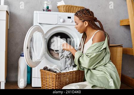 Afroamerikanerin mit Afro-Zöpfen, die an einer Waschmaschine sitzt und im Badezimmer Wäsche macht. Stockfoto