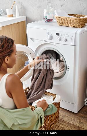 Eine Afroamerikanerin mit Afro-Zöpfen lädt Kleidung vorsichtig in eine Waschmaschine im Badezimmer. Stockfoto