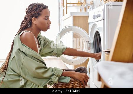 Eine Afroamerikanerin mit Afro-Geflechten lädt eine Waschmaschine in einen Trockner, während sie im Badezimmer Wäsche macht. Stockfoto