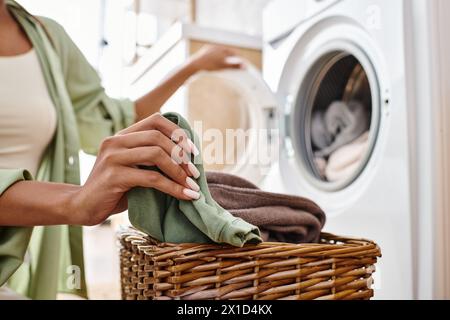 Eine Frau mit Afro-Flechten hält fröhlich einen Wäschekorb neben einer Waschmaschine in einem lebhaften Badezimmer. Stockfoto