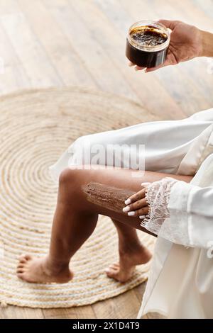 Eine Frau im Bademantel sitzt auf einer Badewanne und peelt das Bein mit Peeling Stockfoto