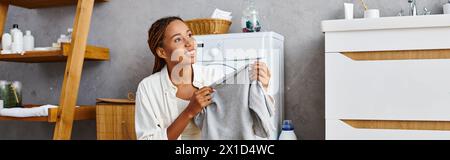 Eine afroamerikanische Frau mit Afro-Flechten hält spielerisch eine Kleidung in der Nähe einer Waschmaschine Stockfoto