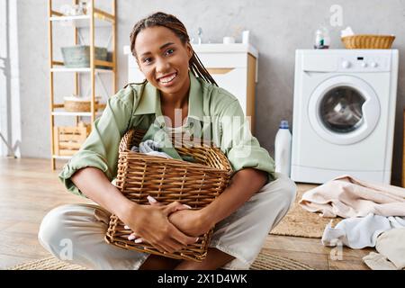 Eine Afroamerikanerin mit Afro-Zöpfen kniet auf dem Boden und hält anmutig einen Korb, während sie im Badezimmer Wäsche macht. Stockfoto