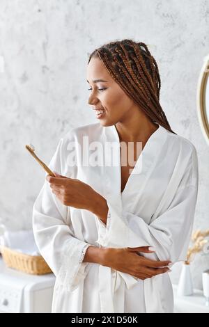 Eine afroamerikanische Frau in einem weißen Gewand hält zart einen Pinsel und strahlt Kreativität und Anmut in einem modernen Badezimmer aus. Stockfoto
