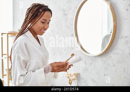 Eine Afroamerikanerin mit Afro-Zöpfen steht in einem modernen Badezimmer und hält einen Pinsel in einem Bademantel. Stockfoto