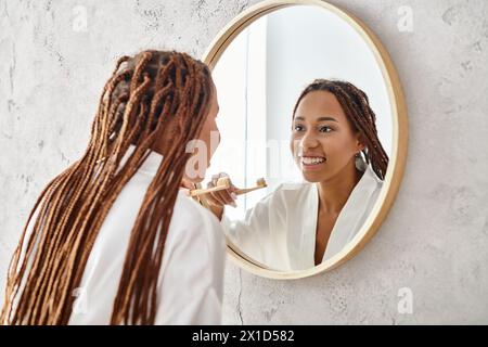 Eine Afroamerikanerin mit Afro-Zöpfen in einem Bademantel, die sich in einem modernen Badezimmerspiegel die Zähne putzt. Stockfoto