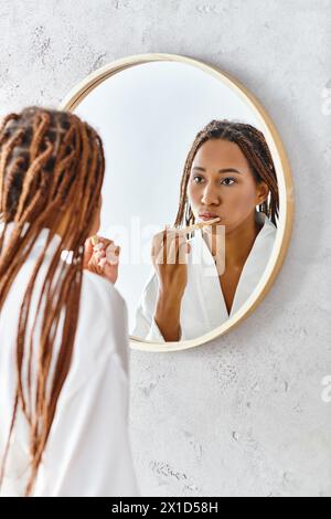 Eine Afroamerikanerin mit Afro-Zöpfen im Bademantel putzt sich die Zähne vor einem Spiegel in einem modernen Badezimmer. Stockfoto