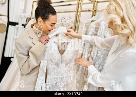 Zwei junge Frauen, eine schöne Braut und ein Ladenassistent, die ein Kleid, das an einem Regal in einer Boutique hängt, sorgfältig untersuchen. Stockfoto