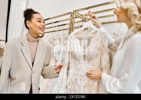 Junge Frauen, die ein Hochzeitskleid auf einem Ständer in einem Brautladen bewundern, unterstützt von einem Ladenleiter. Stockfoto