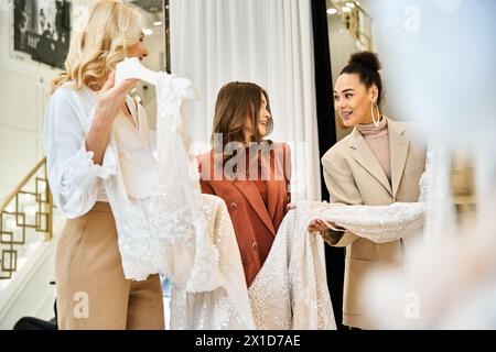 Zwei Frauen, eine Mutter und beste Freundin, bewundern ein wunderschönes Hochzeitskleid auf einer Schaufensterpuppe, während sie mit einer jungen Braut einkaufen. Stockfoto
