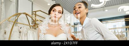Eine junge brünette Braut in einem Hochzeitskleid wird von einem afroamerikanischen Einkaufsassistenten in einem Brautsalon begleitet. Stockfoto