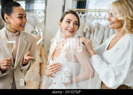 Eine junge brünette Braut in einem Hochzeitskleid lächelt aufgeregt, während ihre Mutter mittleren Alters und beste Freundin, eine Brautjungfer, ihr helfen, einen Schleier anzulegen. Stockfoto