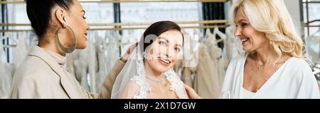 Eine junge brünette Braut, ihre Mutter mittleren Alters und die beste Freundin als Brautjungfer stehen in einem Brautsalon an einem Kleiderständer. Stockfoto