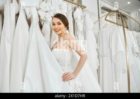 Eine junge brünette Braut steht vor einem Regal mit weißen Kleidern und wählt sorgfältig ihr perfektes Hochzeitskleid aus. Stockfoto