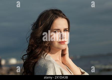 Gesunde Brünette Frau mit frischer Haut, natürlichem Make-up und gesundem welligen braunen Haar genießt die sonnige Morgen- und Meeresluft. Weibliches Modell für den Außenbereich Stockfoto