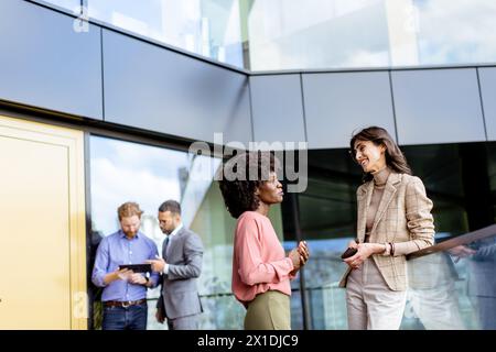 Zwei weibliche multiethnische Kollegen unterhalten sich vor einem Büro freundlich, während andere im Hintergrund arbeiten Stockfoto