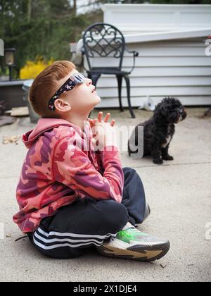 Der 11-jährige Junge sah 2024 Sonnenfinsternis und trug eine Sonnenbrille zum Augenschutz. Hund im Hintergrund. Stockfoto