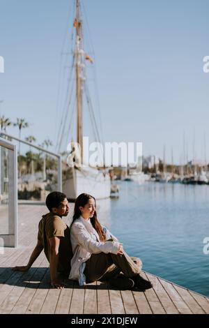 Ein Paar sitzt nahe an einem Dock und genießt einen ruhigen Moment mit Blick auf den Yachthafen an einem sonnigen Tag in Barcelona. Stockfoto