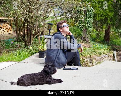 Glücklicher 15-jähriger Junge, der 2024 Sonnenfinsternis beobachtet und Sonnenbrille zum Augenschutz trägt. Hund im Vordergrund. Stockfoto