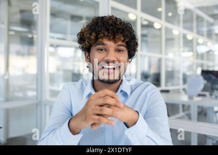 Ein junger männlicher Profi mit lockigem Haar strahlt Selbstvertrauen und Glück aus, während er in einer hellen, modernen Büroumgebung sitzt. Stockfoto
