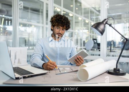 Fokussierter junger, männlicher Architekt mit lockigem Haar, der in einem hellen, modernen Büro mit digitalem Tablet und Laptop an architektonischen Plänen arbeitet. Stockfoto