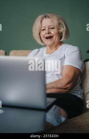 Ältere lächelnde Frau, die einen Laptop für Videoanrufe nutzt oder online mit fröhlicher Stimmung auf dem Sofa zu Hause einkaufen möchte. Stockfoto
