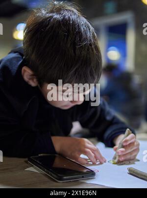 latino kleiner Junge, Linkshänder in einer Bar, unterhielt sich mit Papier und Buntstiften, zeichnete und ohne Bildschirme. Selektiver Fokus Stockfoto