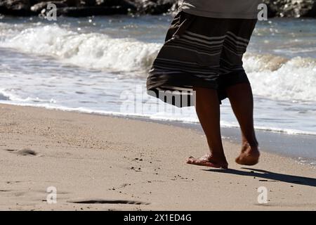 Die untere Hälfte der Frau in einem Kleid von hinten geht barfuß an einem Sandstrand Stockfoto